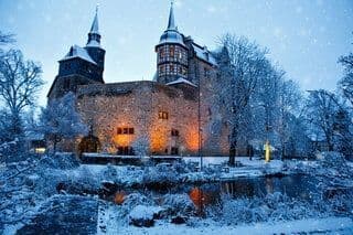 Un castillo de piedra iluminado con dos torres se alza en un paisaje nevado mientras cae la nieve al anochecer.