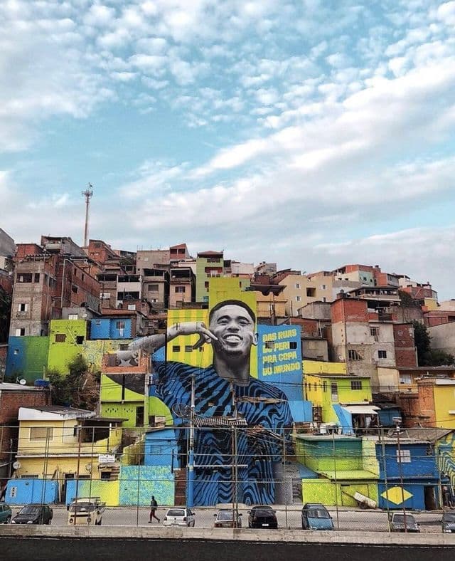 Un grande murale di un uomo sorridente dipinto su diversi edifici colorati su una collina densamente popolata sotto un cielo nuvoloso.