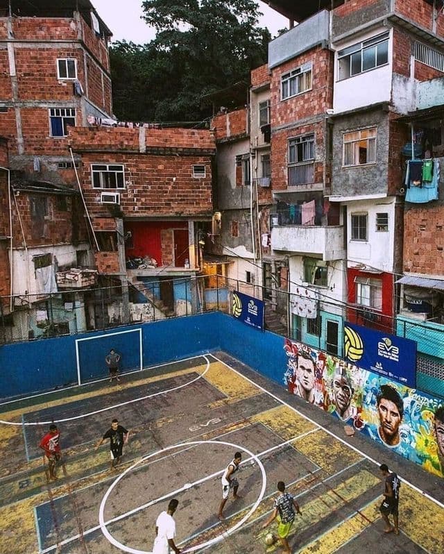 Veduta dall'alto di persone che giocano a calcio su un campo in cemento, circondato da edifici in mattoni fittamente disposti.