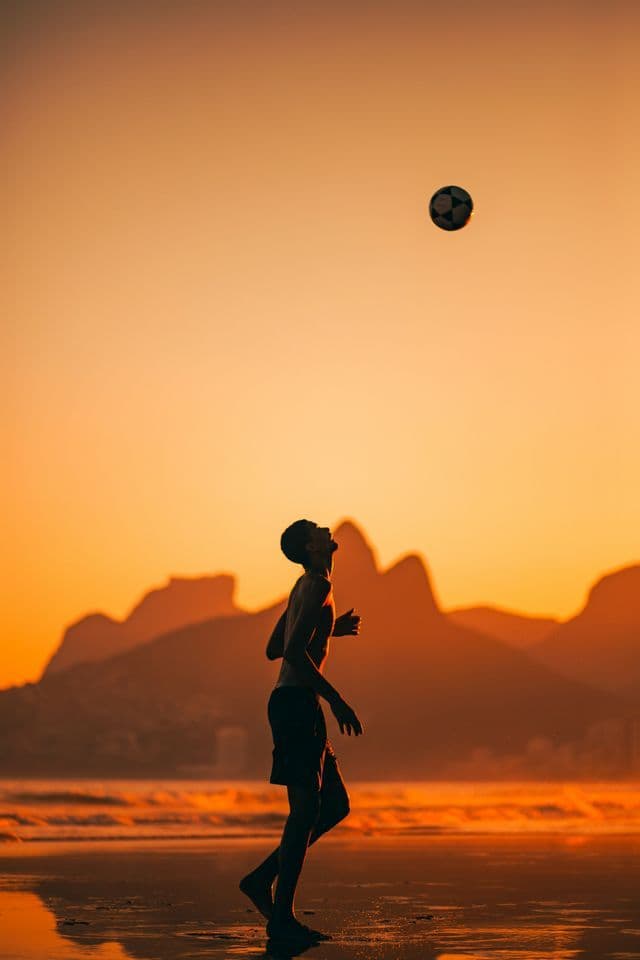 Una persona in silhouette contro un cielo arancione al tramonto gioca con un pallone da calcio su una spiaggia con montagne sullo sfondo.