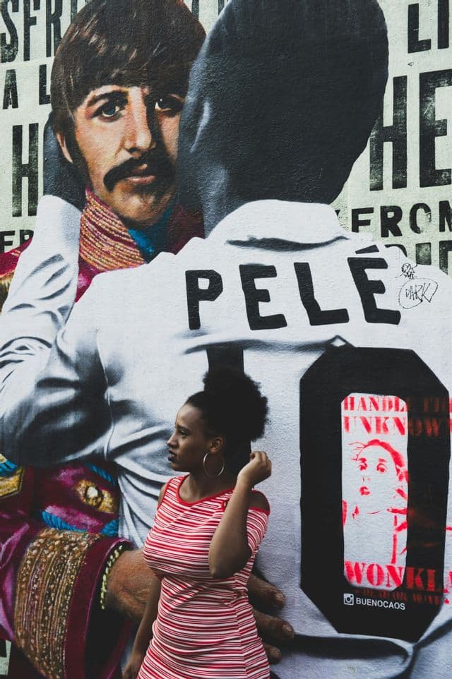 Una donna con un vestito a righe in piedi di profilo davanti a un grande murale di Pelé che abbraccia Ringo Starr.