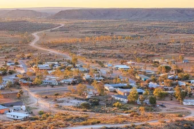 Eine Luftaufnahme einer abgelegenen Ortschaft in einer trockenen Landschaft mit Schotterstraßen und vereinzelten Bäumen während eines goldenen Sonnenuntergangs.