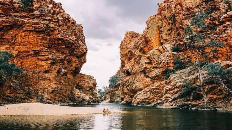 Zwei Teilnehmer einer WeRoad-Gruppenreise sitzen auf einer Sandbank in einer Flussschlucht, umgeben von großen, roten Felsformationen.