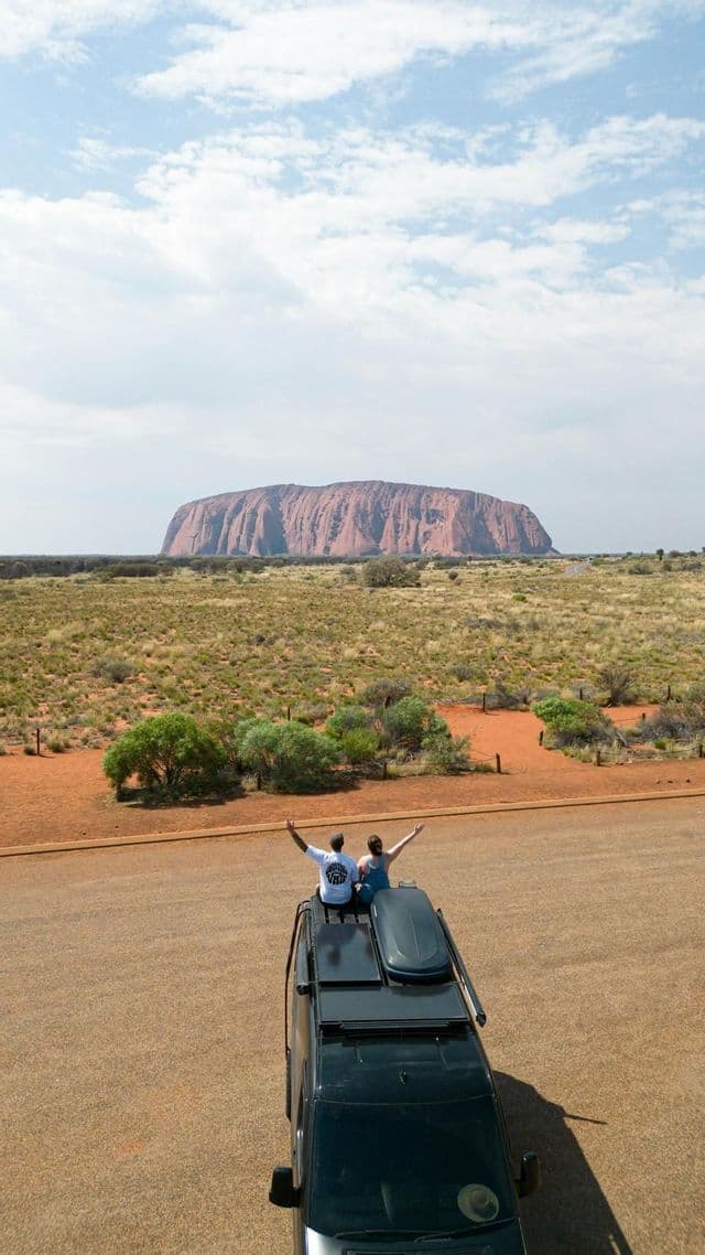 Zwei Personen einer WeRoad-Gruppenreise sitzen mit erhobenen Armen auf einem Fahrzeug und blicken auf einen großen roten Felsmonolithen in einer trockenen Landschaft.
