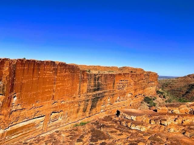 Ein großer oranger Felsencanyon mit steilen, geschichteten Klippen ist unter einem klaren, tiefblauen Himmel in einer sonnigen Landschaft zu sehen.