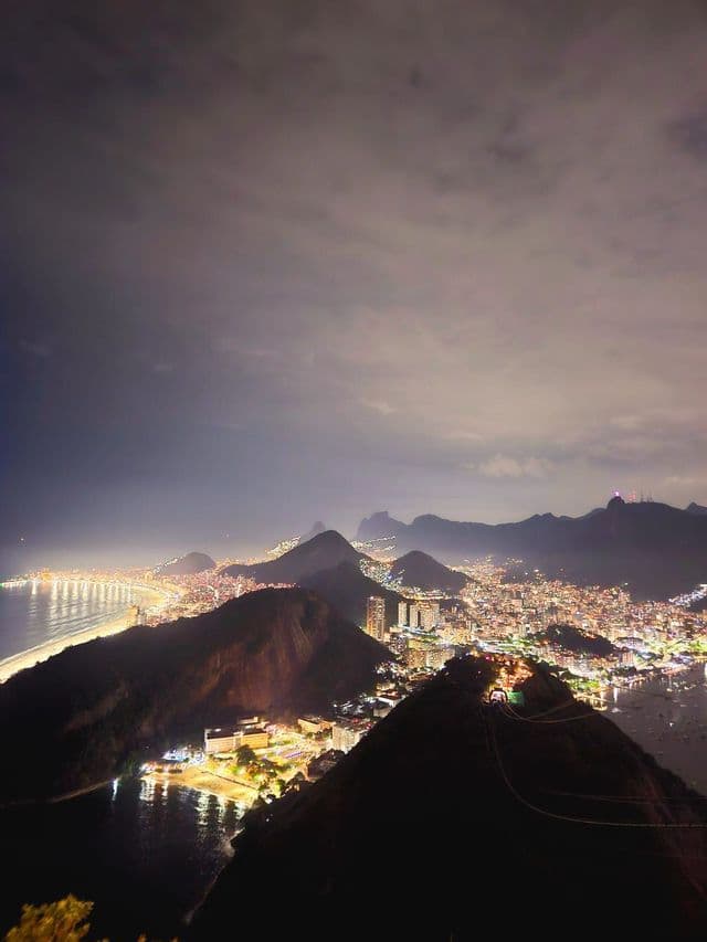 Una vista panorámica de una ciudad costera iluminada por la noche, anidada entre montañas oscuras bajo un cielo nublado.