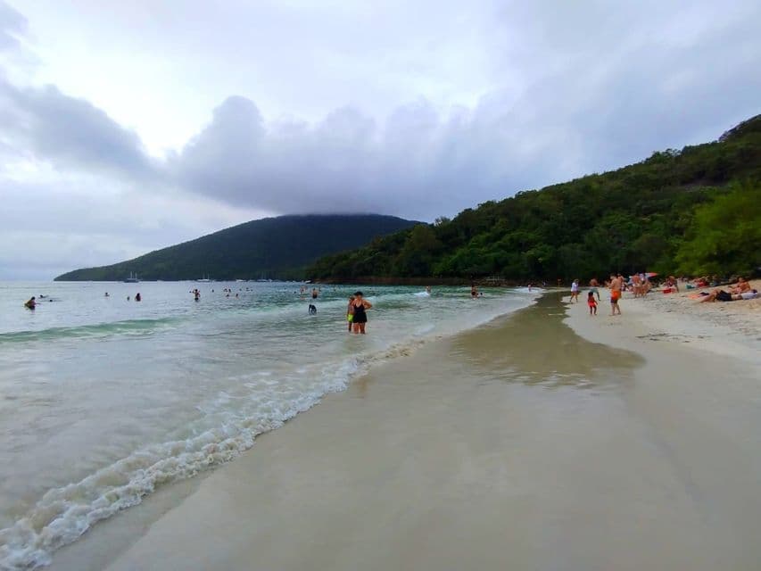 Personas nadan y se relajan en una playa de arena blanca junto a una colina boscosa bajo un cielo nublado.