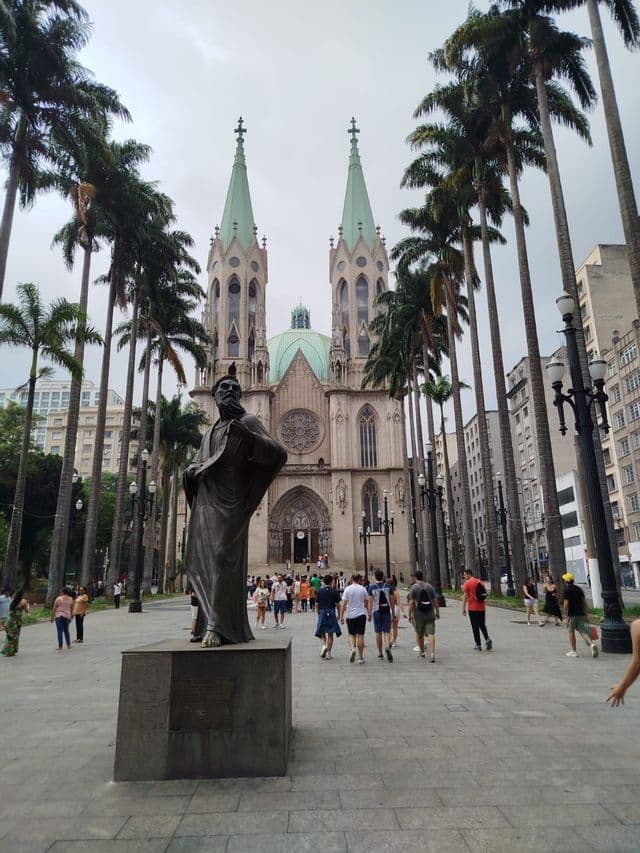 Una plaza amplia con una estatua de bronce y altas palmeras que conduce a una gran catedral con dos agujas de cúpula verde bajo un cielo nublado.