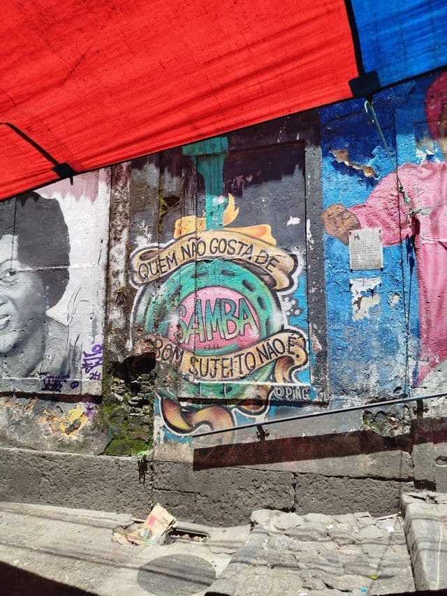 Grafitis coloridos, incluyendo texto sobre samba, pintados en un muro de hormigón desgastado bajo una lona roja.