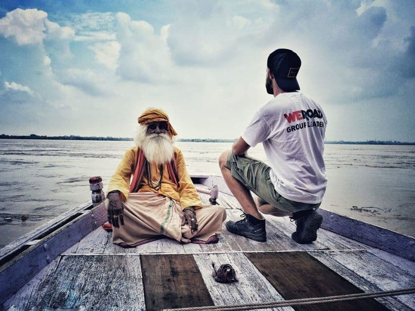 Un líder de grupo WeRoad se sienta con un hombre mayor con una barba blanca larga en la proa de un barco de madera en un río.