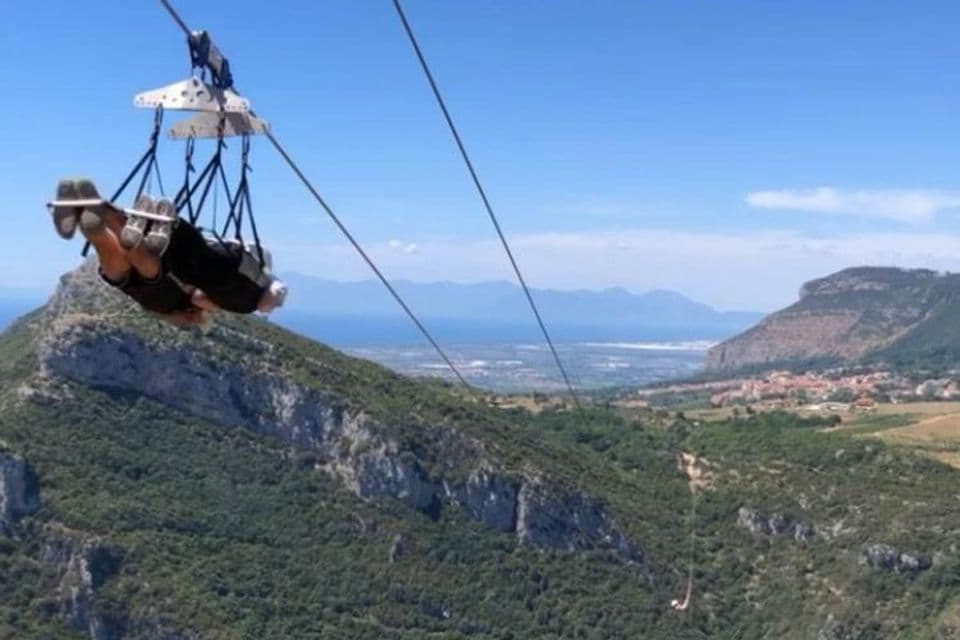 Una persona en arnés vuela de cabeza en tirolina sobre un valle verde y montañoso con el mar a lo lejos.