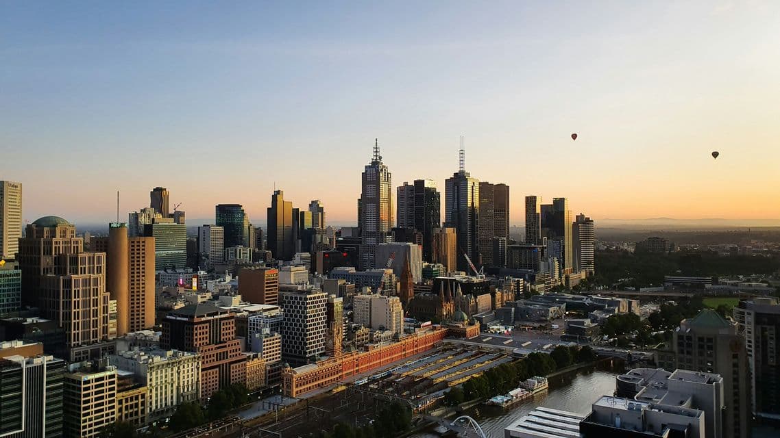 Luftaufnahme einer dichten Stadtlandschaft mit Wolkenkratzern am Fluss bei Sonnenaufgang und zwei Heißluftballons am Himmel.