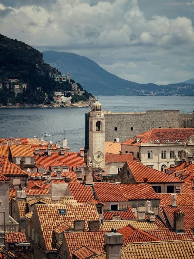 Ein steinerner Uhrturm erhebt sich über roten Ziegeldächern in einer Küstenstadt, mit dem Meer und Bergen im Hintergrund.