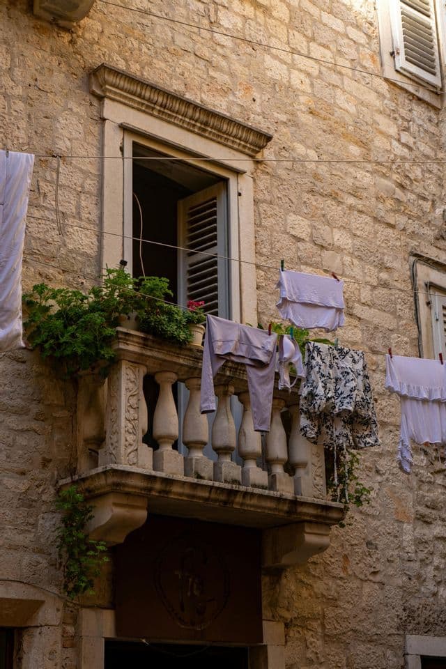 Wäsche trocknet an einer Wäscheleine vor einem verzierten Steinbalkon mit offenem Fensterladen.