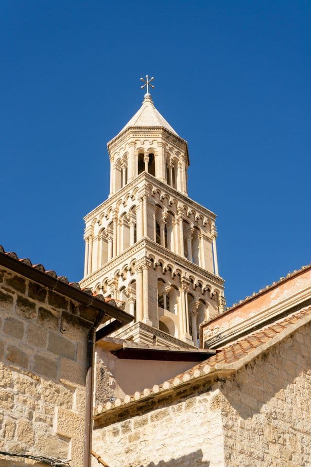 Ein verzierter steinerner Glockenturm mit Kreuz auf der Spitze, über Ziegeldächern vor klarem blauem Himmel gesehen.