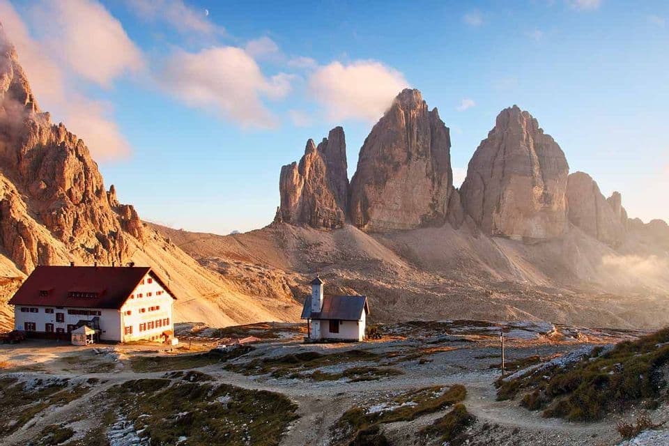 Eine Berghütte und eine kleine Kapelle liegen auf einem felsigen Plateau am Fuße zerklüfteter Gipfel unter blauem Himmel mit einer Mondsichel.