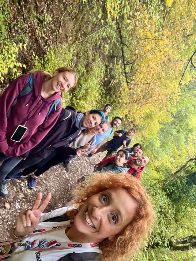 Una donna scatta un selfie sorridente con il suo gruppo di viaggio WeRoad su un sentiero sterrato circondata da alberi verdi e lussureggianti.