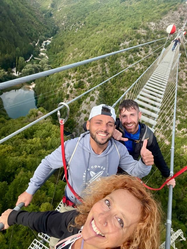 Un gruppo WeRoad sorride per un selfie mentre attraversa un alto ponte sospeso sopra una lussureggiante valle verde e un fiume.