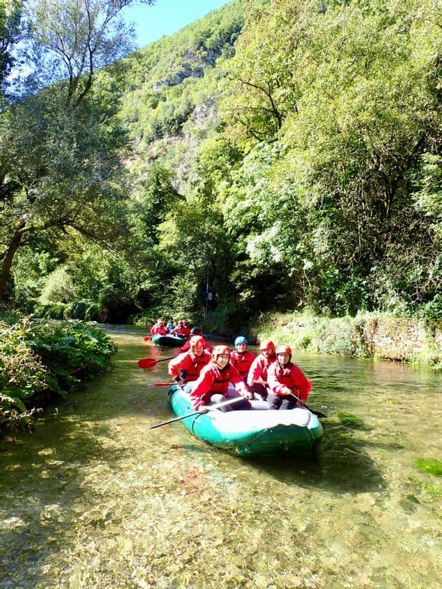 Un viaggio di gruppo WeRoad, con giubbotti di salvataggio rossi, che rema con gommoni gonfiabili lungo un fiume cristallino circondato da alberi verdi lussureggianti.