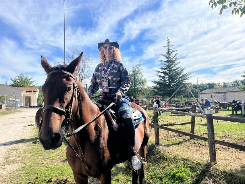Una donna con cappello da cowboy sorride alla macchina fotografica mentre cavalca un cavallo marrone in campagna tra gli alberi.
