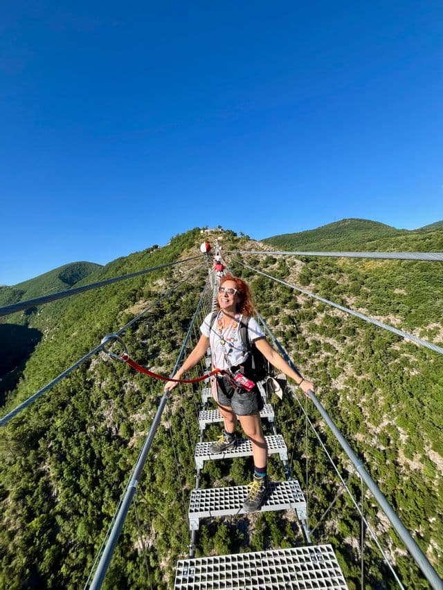 Una donna con i capelli rossi in un viaggio di gruppo WeRoad sorride mentre si trova su un alto ponte sospeso sopra una valle verde e boscosa.