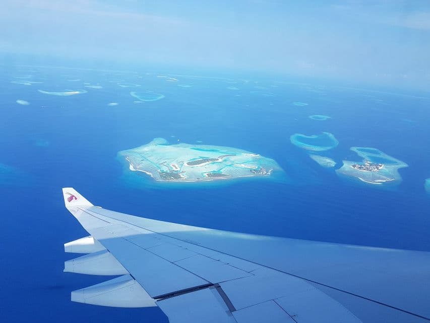 Vue aérienne d'un avion avec son aile au-dessus d'atolls tropicaux dispersés dans un vaste océan bleu profond.