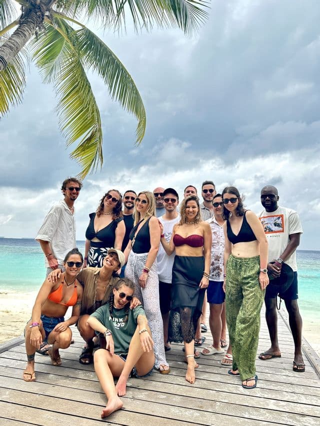 Un groupe WeRoad pose pour une photo sur un ponton en bois, au bord d'une eau turquoise, sous un palmier.