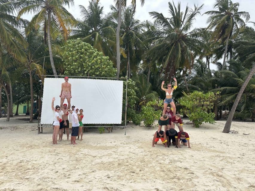 Un voyage de groupe WeRoad forme deux pyramides humaines sur une plage de sable avec des palmiers en arrière-plan.