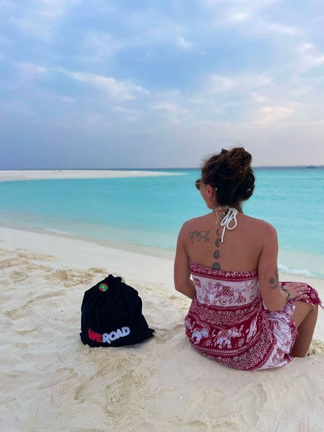 Une femme vue de dos est assise sur une plage de sable blanc à côté d'un sac WeRoad, regardant l'eau turquoise.