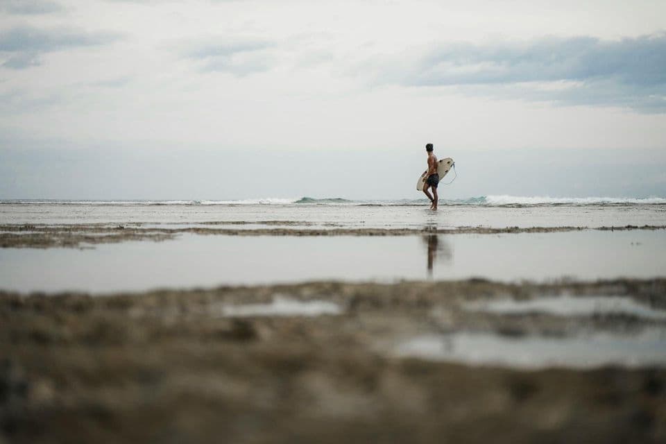 Un hombre lleva una tabla de surf mientras camina por aguas poco profundas del océano en una playa bajo un cielo nublado.