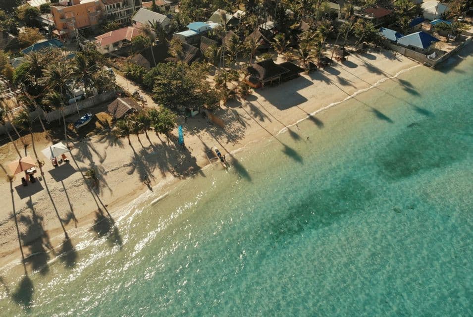 Una vista aérea de una playa tropical con agua cristalina color turquesa, donde las palmeras proyectan largas sombras sobre la arena blanca.