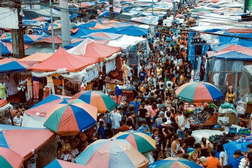 Vista aérea de una gran multitud de personas caminando por un mercado al aire libre, lleno de sombrillas y puestos coloridos.