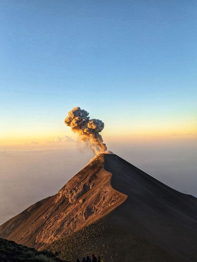 Un volcán entra en erupción con una gran columna de humo al amanecer mientras un viaje en grupo de WeRoad observa desde una cresta.
