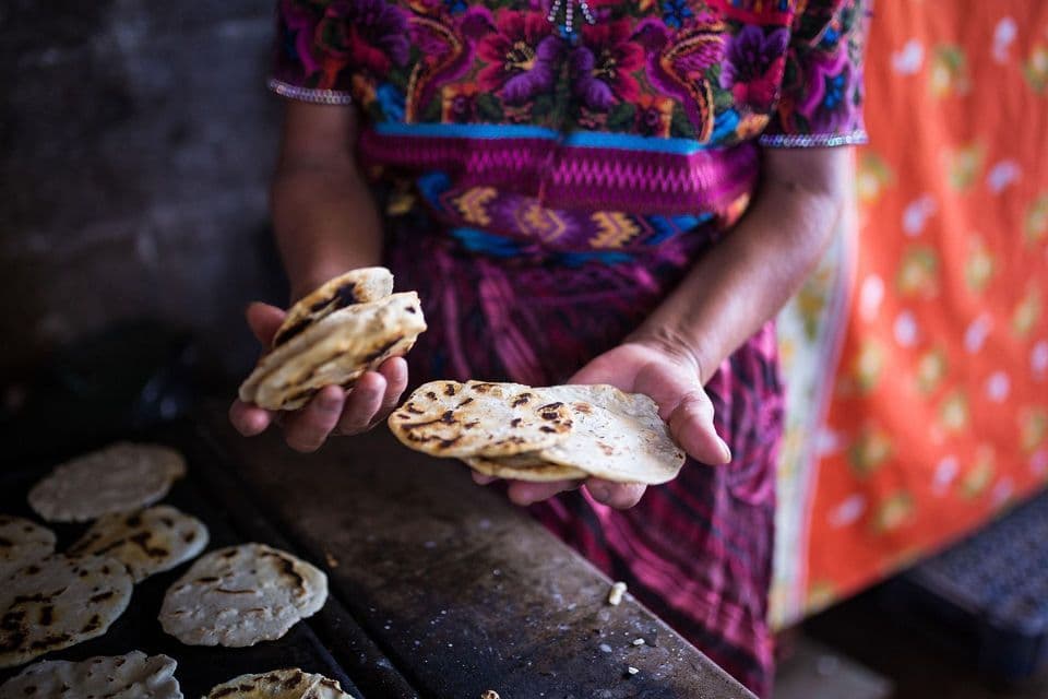 Una persona con una blusa colorida sostiene panes planos recién asados sobre una plancha donde se están cocinando más.