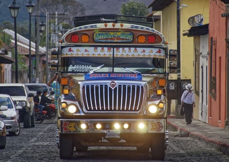 Un autobús brillantemente decorado con las luces encendidas transita por una estrecha calle empedrada bordeada de edificios coloridos.