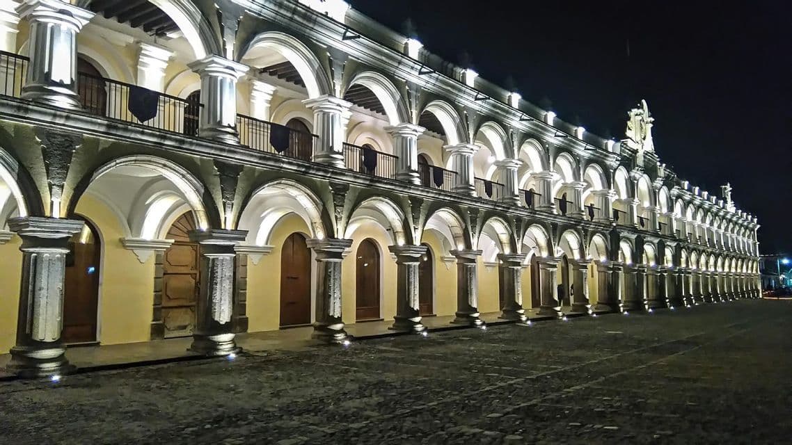 Un largo edificio colonial con una arcada iluminada de dos pisos de arcos y pilares junto a una calle empedrada por la noche.