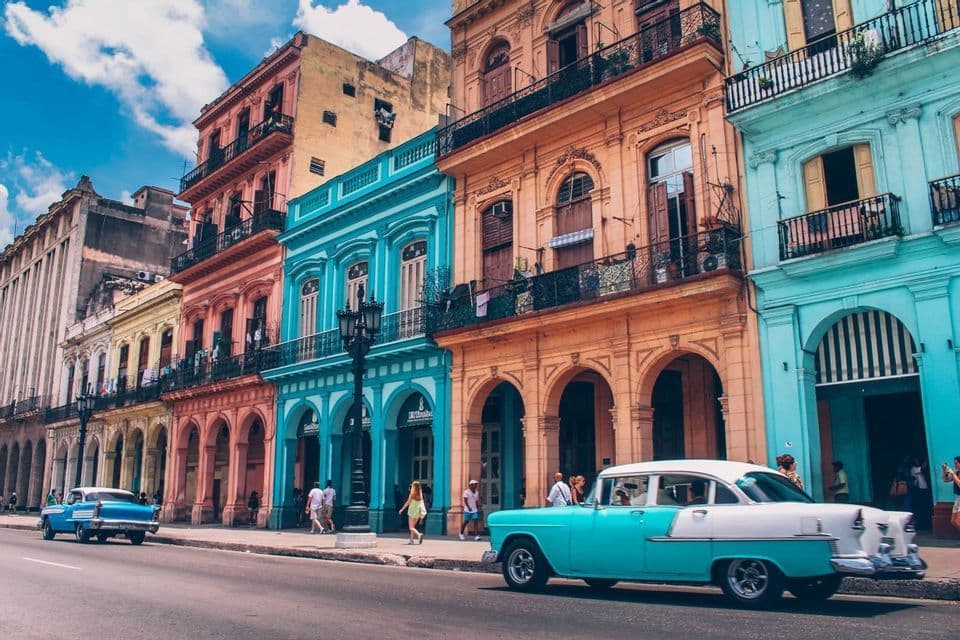 A turquoise and white vintage car is parked on a street next to a row of colorful, ornate buildings under a blue sky.