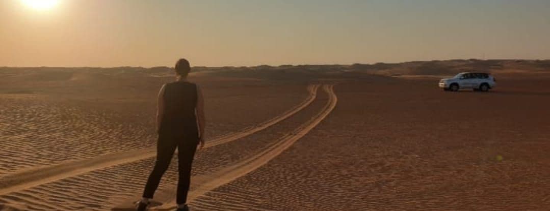 Eine Person steht von hinten in einer Wüstenlandschaft bei Sonnenuntergang, mit Reifenspuren und einem weißen SUV in der Ferne.