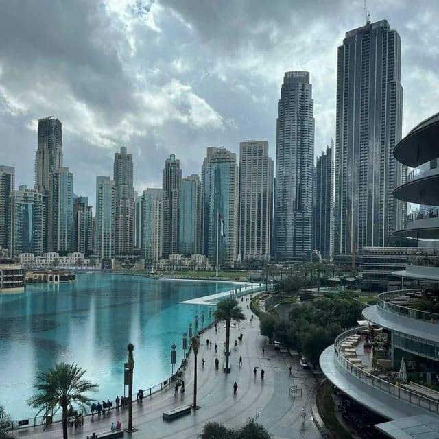 Ein erhöhter Blick auf eine moderne Stadtlandschaft mit Wolkenkratzern, die einen türkisfarbenen See und eine Promenade umgeben, auf der Menschen spazieren gehen.