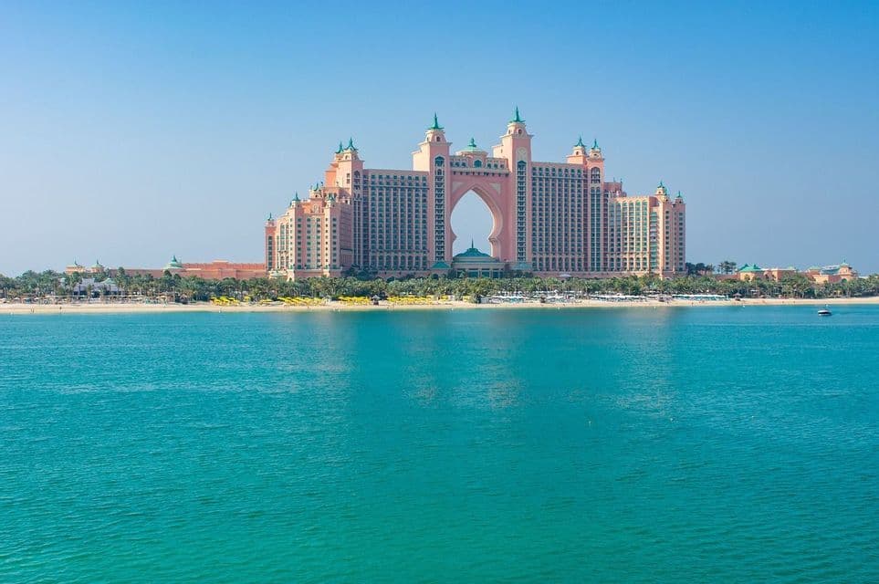 Ein großes, rosafarbenes Hotel mit einem zentralen Bogen steht an einem Sandstrand, von türkisfarbenem Wasser unter klarem blauem Himmel aus betrachtet.