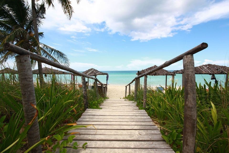 A wooden boardwalk leads through lush green grass down to a sandy beach with turquoise water and thatched-roof huts.
