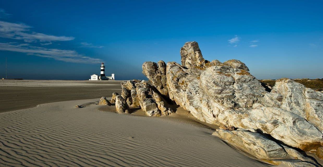 Eine große Felsformation liegt an einem gewellten Sandstrand, mit einem schwarz-weißen Leuchtturm in der Ferne unter einem blauen Himmel.