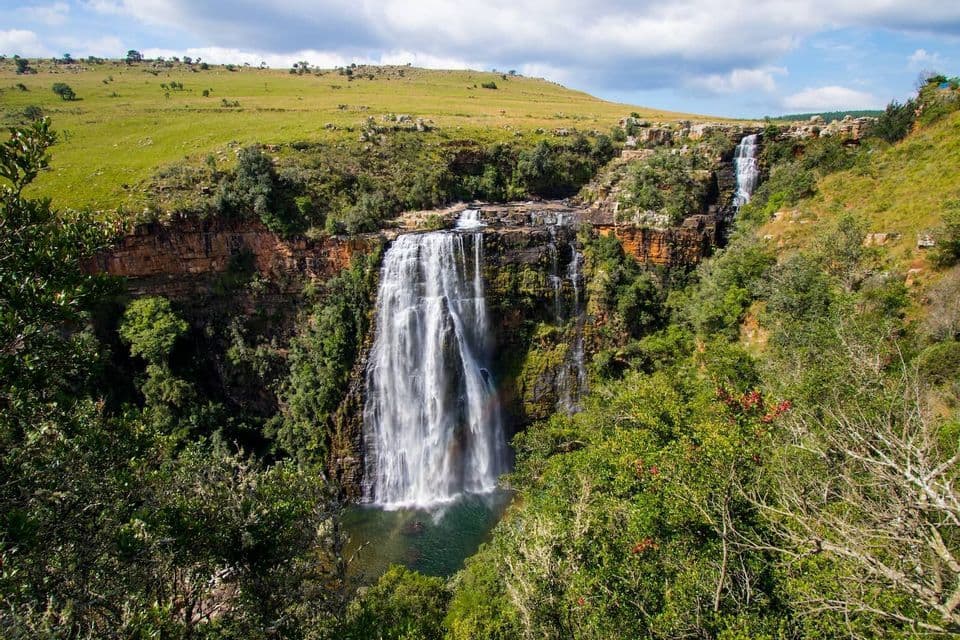 Ein großer Wasserfall stürzt eine felsige Klippe hinab in ein Tauchbecken, umgeben von üppig grünen Bäumen und sanften Hügeln unter einem bewölkten Himmel.