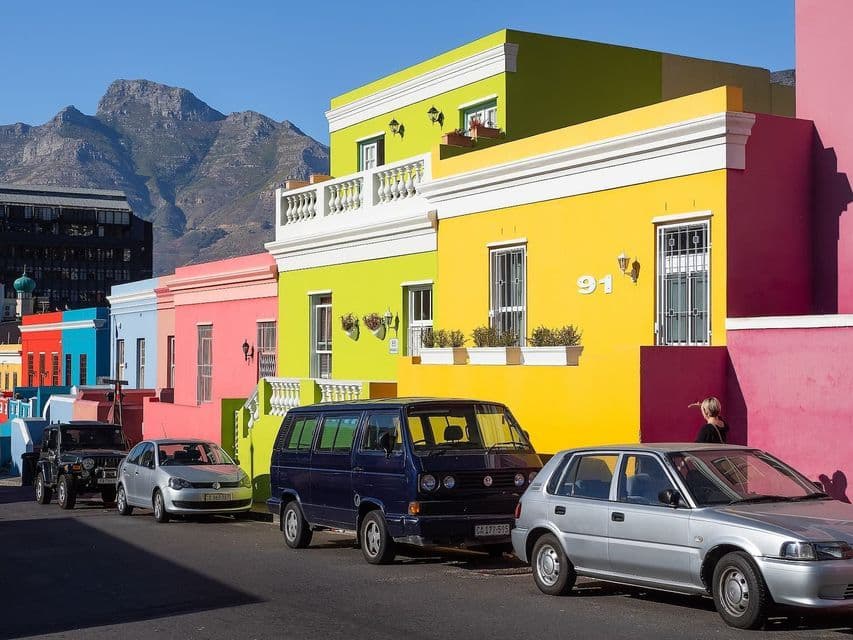 Auto parcheggiate su una strada fiancheggiata da edifici vivaci e colorati, di fronte a una grande montagna sotto un cielo azzurro limpido.