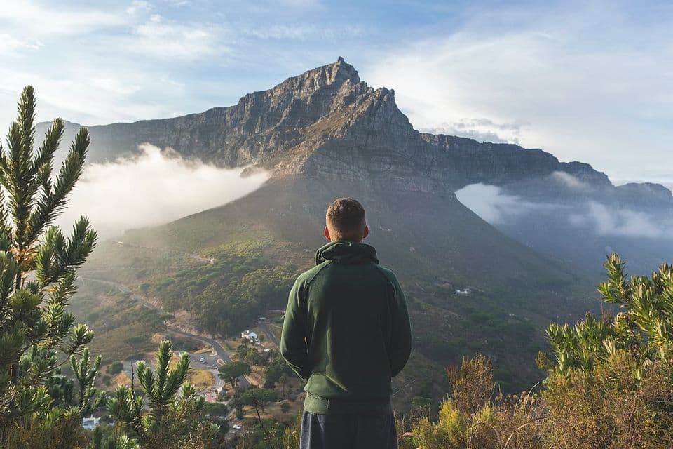 Eine Person in einem grünen Kapuzenpullover steht auf einem Aussichtspunkt und blickt auf einen großen, felsigen Berggipfel, der teilweise von Wolken bedeckt ist.