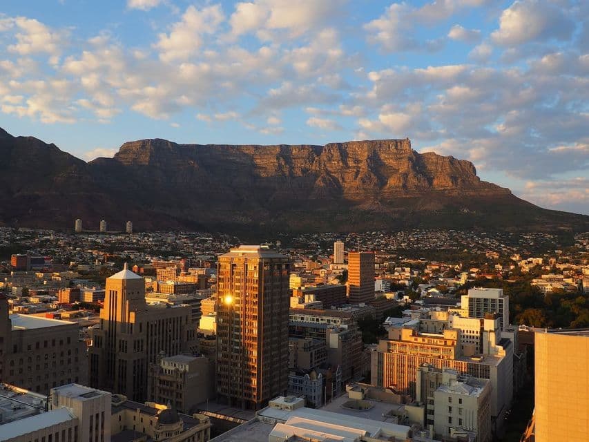 A cityscape with skyscrapers illuminated by golden sunset light, with a large flat-topped mountain in the background under a cloudy sky.