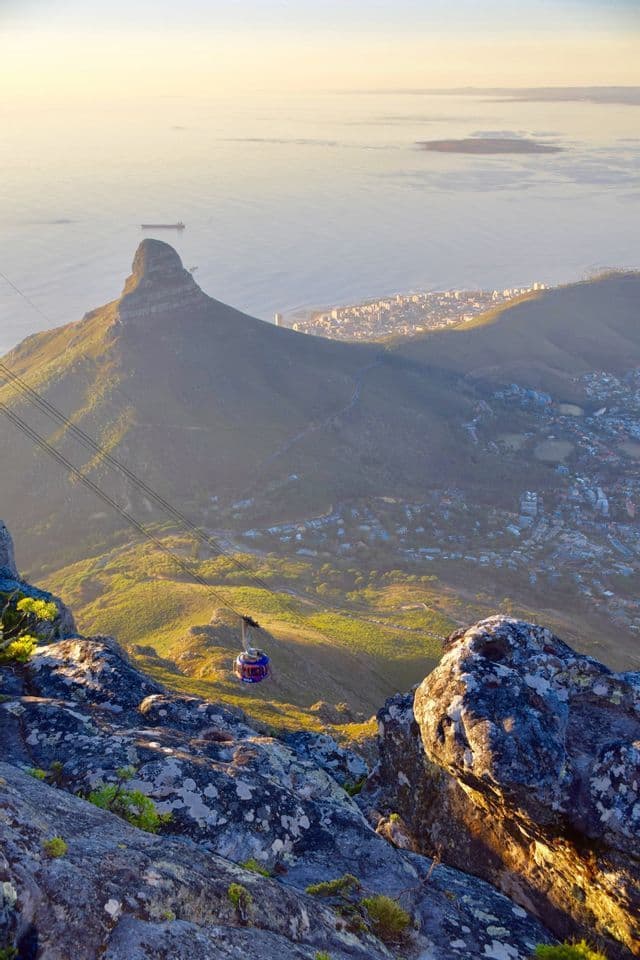 A cable car descends a mountainside, offering a panoramic view of another peak, a coastal city, and the sea during a golden sunrise.