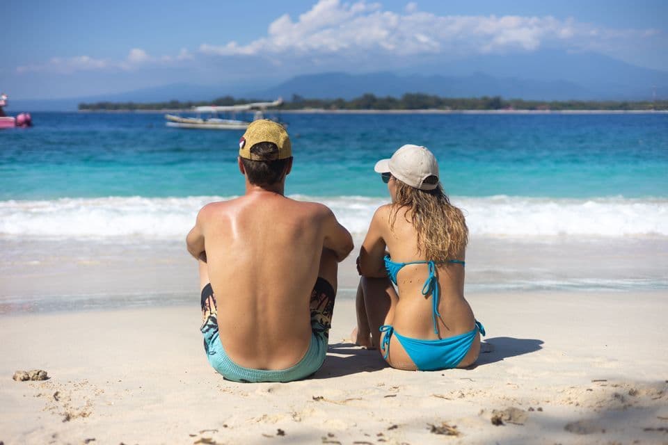 A man and a woman sit on a white sand beach, looking out at the turquoise ocean and distant mountains on a sunny day.