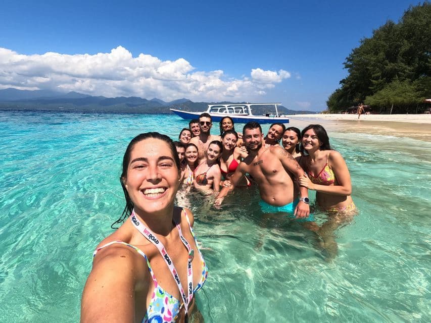 A woman takes a selfie of her WeRoad group trip as they smile together in the clear turquoise water of a tropical beach.