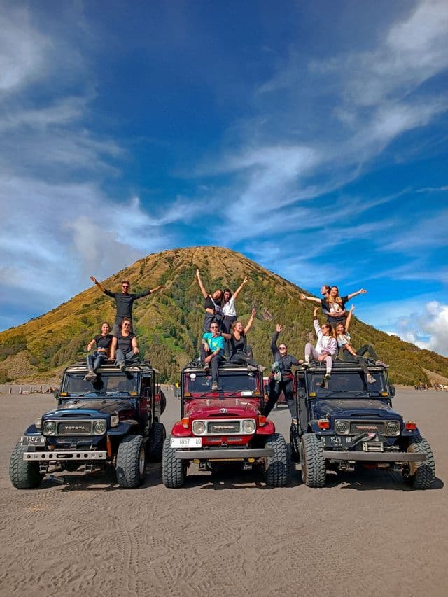 Un gruppo di viaggio WeRoad posa su tre veicoli fuoristrada di fronte a una grande montagna erbosa sotto un cielo parzialmente nuvoloso.